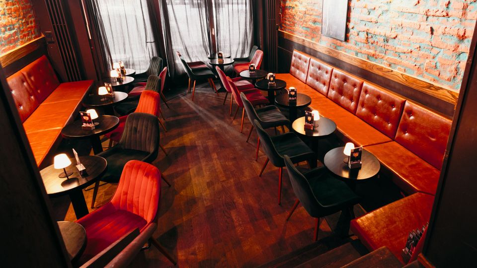 Innenraum einer Bar oder Lounge mit langen, braunen Leder-Sitzbänken entlang freiliegender Ziegelwände und mehreren kleinen runden Tischen mit Tischlampen. Dunkle Polsterstühle, Holzboden und große Fenster mit Vorhängen prägen den schmalen, länglichen Raum.