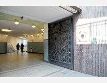 Eingangsbereich einer U-Bahn-Station mit gewölbten Decken, Hängeleuchten und blau gefliesten Wänden, rechts eine verzierte schmiedeeiserne Tür. Zwei Personen gehen in Richtung eines gut beleuchteten Ausgangs.