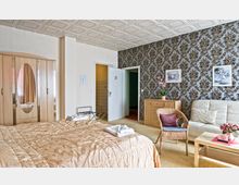 Schlafzimmer mit einem großen beigen Bett, einer Holzgarderobe mit ovalem Spiegel, einem weiß-beige gemusterten Sofa, einem kleinen Holztisch und einer mit Blumen dekorierten Kommode. Die Wand ist mit dunkelgemustertem Tapetendesign verziert, und im Hintergrund sind zwei Türen zu sehen.