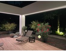 Überdachte Terrasse bei Nacht mit Holzdeck, weißer Stütze und integrierten LED-Lichtleisten entlang der Decke. Im Vordergrund stehen eine Liege und ein kleiner Beistelltisch, umgeben von großen Pflanzkübeln mit Bambus und blühenden Sträuchern sowie Gartenbeleuchtung im Hintergrund.