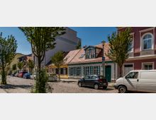 Verkehrsberuhigte Straße mit Kopfsteinpflaster, gesäumt von Bäumen und geparkten Autos. Links und rechts stehen farbenfrohe, historische Wohnhäuser mit Satteldächern und teilweise dekorativen Fensterläden.