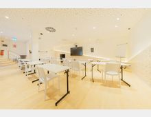 Heller Seminarraum mit weißer Einrichtung, bestehend aus rechteckigen Tischen und Stühlen, auf hellem Holzfußboden. An der Wand befinden sich eine Leinwand, ein Flachbildschirm und ein Flipchart, während die perforierte Decke mit eingelassenen Leuchten ausgestattet ist.