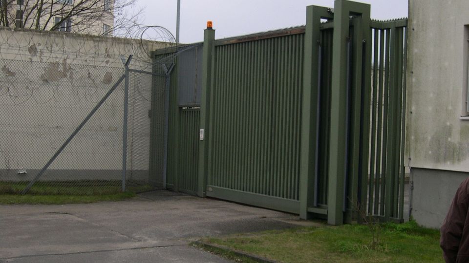 Großes grünes Metallschiebetor neben einer grauen Betonmauer und einem Maschendrahtzaun mit Stacheldraht, davor eine asphaltierte Zufahrt. Im Hintergrund stehen ein hoher Straßenmast, kahle Bäume und ein mehrstöckiges Gebäude.