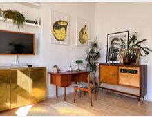 Wohnzimmer mit Retro-Einrichtung: Ein Raum mit einem Mid-Century-Holzschreibtisch, einem kleinen Retro-Stuhl, einem Vintage-Sideboard mit alter Radioeinheit, Zimmerpflanzen und Wandbildern. Links ein goldener Schrank mit einem Flachbildfernseher darüber, auf hellem Holzfußboden.