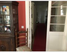 Wohnraum mit antikem Holzschrank und verzierter Holzstuhllehne; rechts eine weiße Glastür, die zu einem langen, roten Teppichläufer in einem Flur führt. Die Wand ist rot gestrichen, und im Hintergrund sind Türen und Möbel sichtbar.