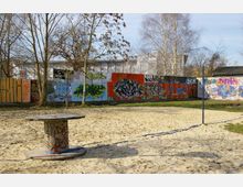 Ein sandiger Außenbereich mit einem großen Holzkabeltrommel-Tisch im Mittelpunkt. Im Hintergrund steht eine bunt bemalte Wand mit Graffiti, umgeben von kahlen Bäumen und vereinzelten Grasflächen.