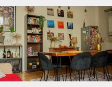 Eclectic dining room with a large wooden table and black upholstered chairs beneath hanging pendant bulbs. The space features a tall bookcase, a wall of framed art, a sideboard with decor, and a sticker-covered cabinet, with plants and a small bar shelf along the side.