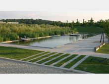 Ein Naturbad mit rechteckigem Becken, umgeben von Schilf und Rasenflächen. Im Vordergrund sind gepflasterte Wege und Liegen zu sehen; im Hintergrund eine bewaldete Landschaft.