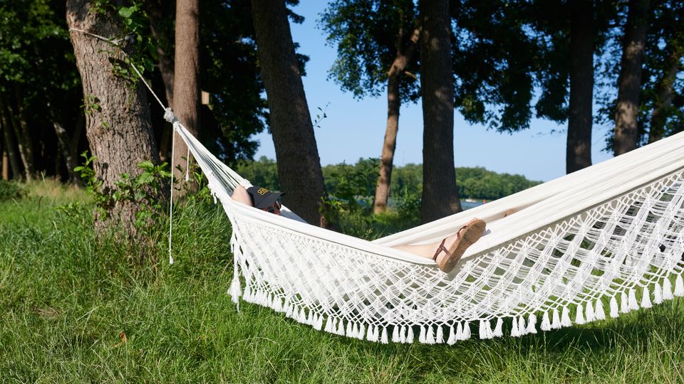 Eine weiße Hängematte mit Fransen ist zwischen zwei Bäumen in einem grasbewachsenen Waldgebiet aufgehängt, darin liegt eine Person mit Sonnenhut. Im Hintergrund sind weitere Bäume und ein blauer Himmel zu sehen.