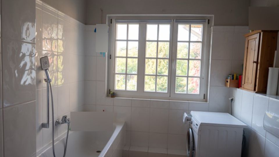Helles Badezimmer mit weiß gefliesten Wänden, Badewanne mit Handbrause links und einem großen, sprossigen Fenster in der Mitte. Rechts stehen eine Waschmaschine unter einer Ablage sowie ein hölzerner Wandschrank; der Boden ist gefliest.