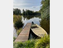 Ein Holzsteg führt zu einem kleinen Motorboot, das in einem ruhigen See verankert ist. Der See ist von Bäumen und Schilf umgeben, und die Szene wird von Sonnenschein und einem leicht bewölkten Himmel beleuchtet.