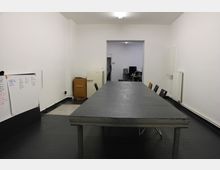 Ein einfacher Besprechungsraum mit einem langen, dunklen Tisch und mehreren Stühlen. An der linken Wand befinden sich ein Whiteboard mit handschriftlichen Notizen, ein kleiner Schrank und ein Kühlschrank, während der Durchgang im Hintergrund in einen weiteren Raum mit Büroausstattung führt.