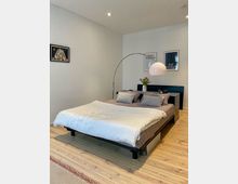 Minimalistisch eingerichtetes Schlafzimmer mit einem Doppelbett, hellem Holzfußboden und weißer Wand. Über dem Bett befinden sich Bilderrahmen, daneben eine gebogene Stehlampe mit rundem Lampenschirm; auf dem Nachttisch steht eine Vase mit Blumen.