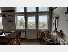 Ein Zimmer mit großen Fenstern und einer Balkontür, durch die eine Ansicht von Häusern und Bäumen sichtbar ist. Im Innenraum befinden sich alte Werkzeuge an den Wänden, ein Tisch mit Utensilien, ein Heizkörper und ein geflochtener Korb auf einer Bank.