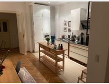 Offene Küche mit weißen Schränken, schwarzer Arbeitsplatte und Holzelementen, darunter ein freistehender Küchentisch mit Ablagefächern. Der Raum hat einen hölzernen Fischgrätenboden und warme Beleuchtung, ergänzt durch Bilder an der Wand und Dekorationen wie Blumenvasen.