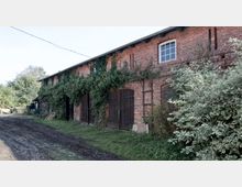 Backsteinbau mit hölzernen Scheunentoren, von Efeu und anderen Pflanzen überwuchert, flankiert von einem großen Busch. Im Vordergrund ein unbefestigter, matschiger Weg, umgeben von Wiese und Bäumen.