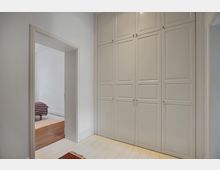 Flur mit weiß gestrichenen Wänden und hellen Holzdielen, ausgestattet mit einem großen, eingebauten Kleiderschrank mit weißen Paneeltüren. Eine offene Tür führt in ein angrenzendes Schlafzimmer mit dunklem Holzboden und einem Bett mit gestreifter Decke.