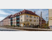 Straßenansicht mit mehrstöckigen Wohngebäuden im Stil der frühen Moderne, gekennzeichnet durch rote Ziegelfassaden, helle Fensterrahmen und Satteldächer. Im Vordergrund verlaufen Straßenbahnschienen und Oberleitungen, daneben ein Gehweg mit einzelnen Bäumen und einem Geländer.