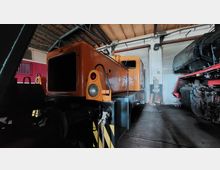 Eine orangefarbene Diesellokomotive steht in einer Halle mit Holzdach und Rolltoren, umgeben von weiteren Zugteilen und technischen Elementen.