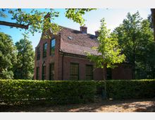 Backsteingebäude mit Satteldach und grünen Fensterrahmen, umgeben von einer niedrigen Hecke und Bäumen in einer grünen Umgebung. Das Gebäude steht bei sonnigem Wetter, und das Gelände zeigt Anzeichen von Herbstlaub am Boden.