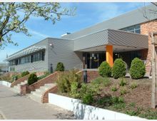 Modernes Gebäude mit Flachdach und metallverkleideter Fassade, kombiniert mit roten Klinkersteinen. Im Vordergrund sind Treppen mit Ziegelsteinseiten sowie bepflanzte Grünflächen und Gehwege zu sehen, während der Eingangsbereich durch eine orange Säule hervorgehoben wird.