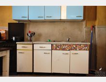 Kleine Küche mit gelben Wänden und einer einfachen Ausstattung. Sichtbar sind eine Spüle mit weißem Unterschrank, teils mit buntem Muster, blaue Hängeschränke, ein schwarzer Herd und einige Küchenutensilien.