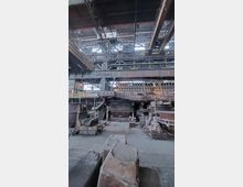 Innenansicht einer stillgelegten Industriehalle mit schweren Maschinen und Stahlstrukturen. Sichtbar sind massive Metallgerüste, Förderanlagen und große industrielle Bauteile, die auf eine ehemalige Nutzung in der Schwerindustrie hinweisen.