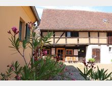 Fachwerkhaus mit hellem Putz, Holzbalken und roten Ziegeln auf dem Dach, umgeben von blühenden Oleanderpflanzen. Im Vordergrund ein Kopfsteinpflasterhof und Holzstühle vor dem Gebäudeeingang.