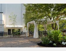 Ein gepflegter Außenbereich mit Kiesboden, einem weißen Pavillon und Gartenmöbeln. Im Hintergrund steht ein helles Gebäude mit Fenstern und einem Baldachin, umgeben von Bäumen und grüner Bepflanzung.