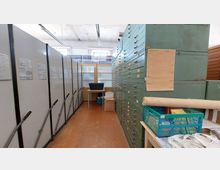 Ein Archivraum mit grauen Schieberegalsystemen auf der linken Seite und hohen, grünen Metallschubladenschränken auf der rechten Seite. Im Hintergrund steht ein kleiner Tisch vor einer Wand mit halb verglasten Türen und Fenstern, durch die Tageslicht einfällt.