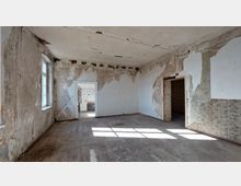 Ein leerer, verfallener Raum in einem alten Gebäude mit abblätterndem Putz an den Wänden, einem Holzboden und sichtbaren Rissen in der Decke. Der Raum hat ein Fenster, durch das Licht einfällt, sowie zwei Türöffnungen, die in angrenzende Räume führen.