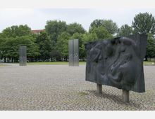 Eine gepflasterte Platzfläche mit einer schwarzen Skulptur im Vordergrund, die Reliefs von menschlichen Figuren zeigt. Im Hintergrund stehen zwei schmale, hohe Metallsäulen vor einer grünen Baumreihe.