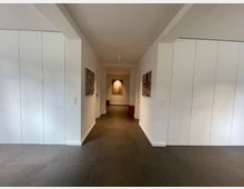 Moderner Flur mit weißen Wänden und großen dunklen Fliesen auf dem Boden, flankiert von Einbauschränken. An den Wänden hängen Kunstwerke, und am Ende des Flurs ist ein weiteres beleuchtetes Bild sichtbar.