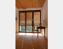 Ein heller Raum mit Holzdecke und Bambusboden, großen Fenstern mit Holzjalousien und einem kleinen Schreibtisch mit Stuhl. Durch die Fenster sind Gitter und grüne Pflanzen im Außenbereich sichtbar.