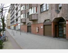Straßenansicht eines mehrstöckigen Wohn- und Geschäftsgebäudes mit grauer Fassade und braunen Klinkerelementen. Der Gehweg ist breit, links stehen Fahrräder und Bäume, rechts sind Eingänge, Fenster und geschlossene Rolltore zu sehen.