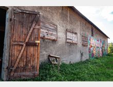 Eine alte Scheune mit grauer Betonwand, auf der bunte Graffiti-Gemälde zu sehen sind. Die Holzfensterläden sind geschlossen, und eine Holztür steht offen; davor wächst hohes Gras.