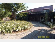 Hof mit Kopfsteinpflasterweg, umgeben von Blumenbeeten mit weißen Rosen und Bäumen. Im Hintergrund ein Gebäude mit Ziegeldach, Holzveranda und rankenden Pflanzen.