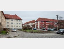 Wohnsiedlung mit mehrstöckigen Gebäuden im typischen Stil der 1920er- bis 1930er-Jahre mit roten Ziegeldächern und weißen oder roten Fassaden. Im Vordergrund eine kleine Grünfläche mit parkenden Autos entlang einer gepflasterten Straße.