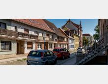 Straßenszene in einer Kleinstadt mit Reihen von zweigeschossigen Häusern mit roten Ziegeldächern und teilweise verputzten Fassaden. Autos parken entlang der gepflasterten Straße, und im Hintergrund ist ein Gebäude mit Ziergiebel und Kirchturm sichtbar.