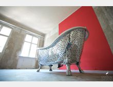 Freistehende Badewanne mit Mosaiksteinchen verziert, auf metallenen Löwenfüßen stehend, in einem industriellen Raum mit unverputzten Wänden, großen Fenstern und einem auffälligen roten Farbakzent.