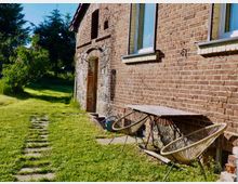 Backsteinfassade eines Landhauses mit zwei Fenstern und einer Holztür, umgeben von einer Wiese mit Gartenmöbeln aus Rattan und einem schmalen Gartenweg aus Steinplatten. Im Hintergrund sind Bäume und Sträucher sichtbar.