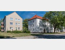 Wohngebiet mit zwei mehrstöckigen Wohngebäuden, eines mit beige Putzfassade und Balkonen, das andere mit weißer Fassade und rotem Satteldach. Vor den Gebäuden befinden sich Rasenflächen, Bäume und ein gepflasterter Gehweg unter blauem Himmel.