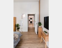 Schlafzimmer mit hellem Holzboden, einem Bett mit grauer Bettdecke und Fellakzent links, gestaltet mit Pflanzen und minimalistischen Kunstwerken an der Wand. Durch eine offene weiße Tür ist eine moderne Küche mit Holz- und weißen Schränken sichtbar.