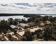 Luftaufnahme einer Landschaft mit einem Gebäudekomplex, bestehend aus mehreren flachen Gebäuden mit hellen Fassaden, umgeben von Bäumen. Im Hintergrund befindet sich ein großer See mit dicht bewaldeten Ufern und vereinzelten Booten auf dem Wasser.