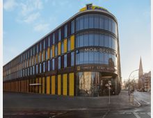 Ein modernes, mehrstöckiges Gebäude mit abgerundeter Ecke und einer Fassade aus Glas, gelben und dunklen Paneelen, beschriftet mit „MOA Berlin“. Im Hintergrund ragt ein Kirchturm in den Himmel, umgeben von Stadtgebäuden.