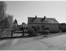 Schwarz-Weiß-Aufnahme eines Einfamilienhauses mit Satteldach und zwei kleinen Schornsteinen, umgeben von einem Garten mit Bäumen und einem Zaun. Im Vordergrund befindet sich eine gepflasterte Einfahrt mit einem geschlossenen Metalltor.