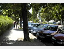 Gehweg in einer von Bäumen gesäumten Straße, links eine Hecke und zwei Personen, die in Richtung des Bildhintergrunds laufen. Rechts parken zahlreiche Autos parallel zur Straße, dahinter sind Wohnhäuser und weitere Bäume zu erkennen.