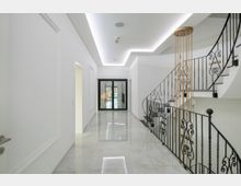 Moderner Flur mit weißen Wänden, glänzendem Marmorboden und einer stilvollen Wendeltreppe mit schwarzem, geschmiedetem Geländer. Am Ende des Flurs befinden sich große Glas-Schiebetüren, die zu einem Außenbereich führen.