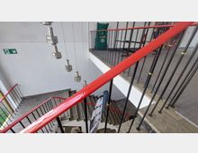 Treppenhaus in einem modernen Gebäude mit roten Handläufen und schwarz-goldenen Geländern. Im Hintergrund hängen geometrische Lampen von der Decke, und ein Notausgangsschild ist an der weißen Wand angebracht.