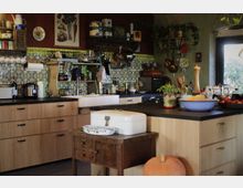 Rustikale Küche mit Holzmöbeln, Fliesen mit buntem Muster an der Wand und schwarzer Arbeitsfläche. Im Raum stehen ein kleiner Holztisch, Dekorationen wie hängende Töpfe, eine große Kürbisfrucht und diverse Küchenutensilien.